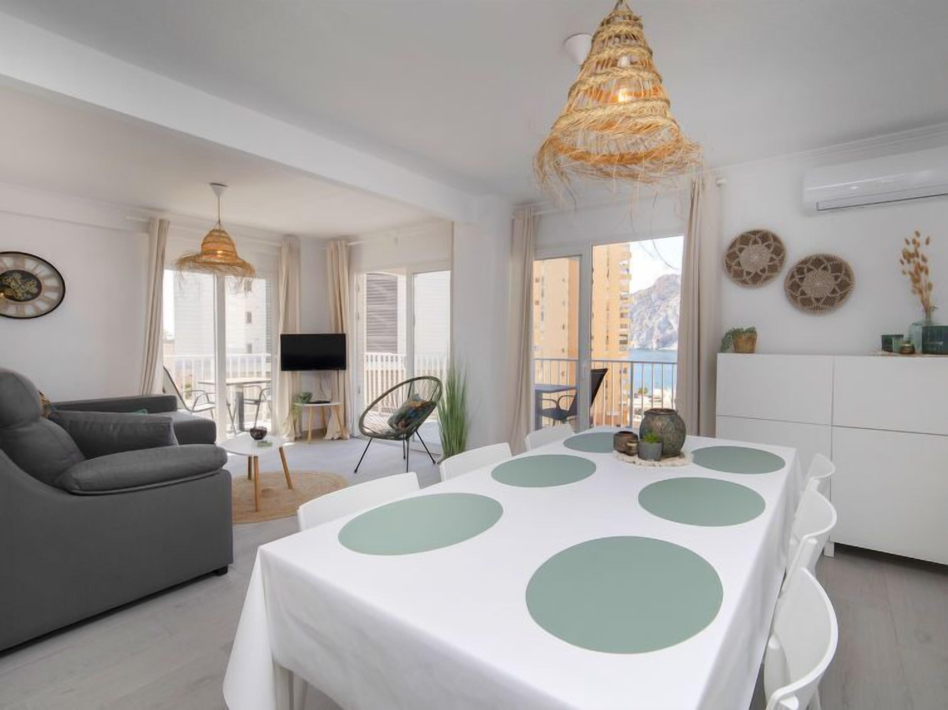 A modern living room with a grey sofa, a dining table with green circular patterns, a hanging light fixture, and a TV on the wall.