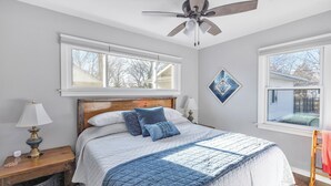Un dormitorio con cama, ventilador de techo, mesita de noche, lámpara y ventana.