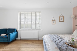 A bedroom with a bed, a blue sofa, a radiator, a window with blinds, and wall-mounted decor. - Coombe Bank (Teignmouth)