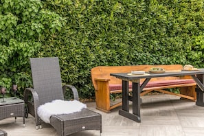 An outdoor seating area with wicker furniture and a wooden bench, set against a backdrop of dense green foliage. - Coombe Bank (Teignmouth)