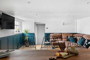 A modern living room with a sofa, chairs, a dining table with a board of cheese and grapes, and a flat-screen TV mounted on the wall.