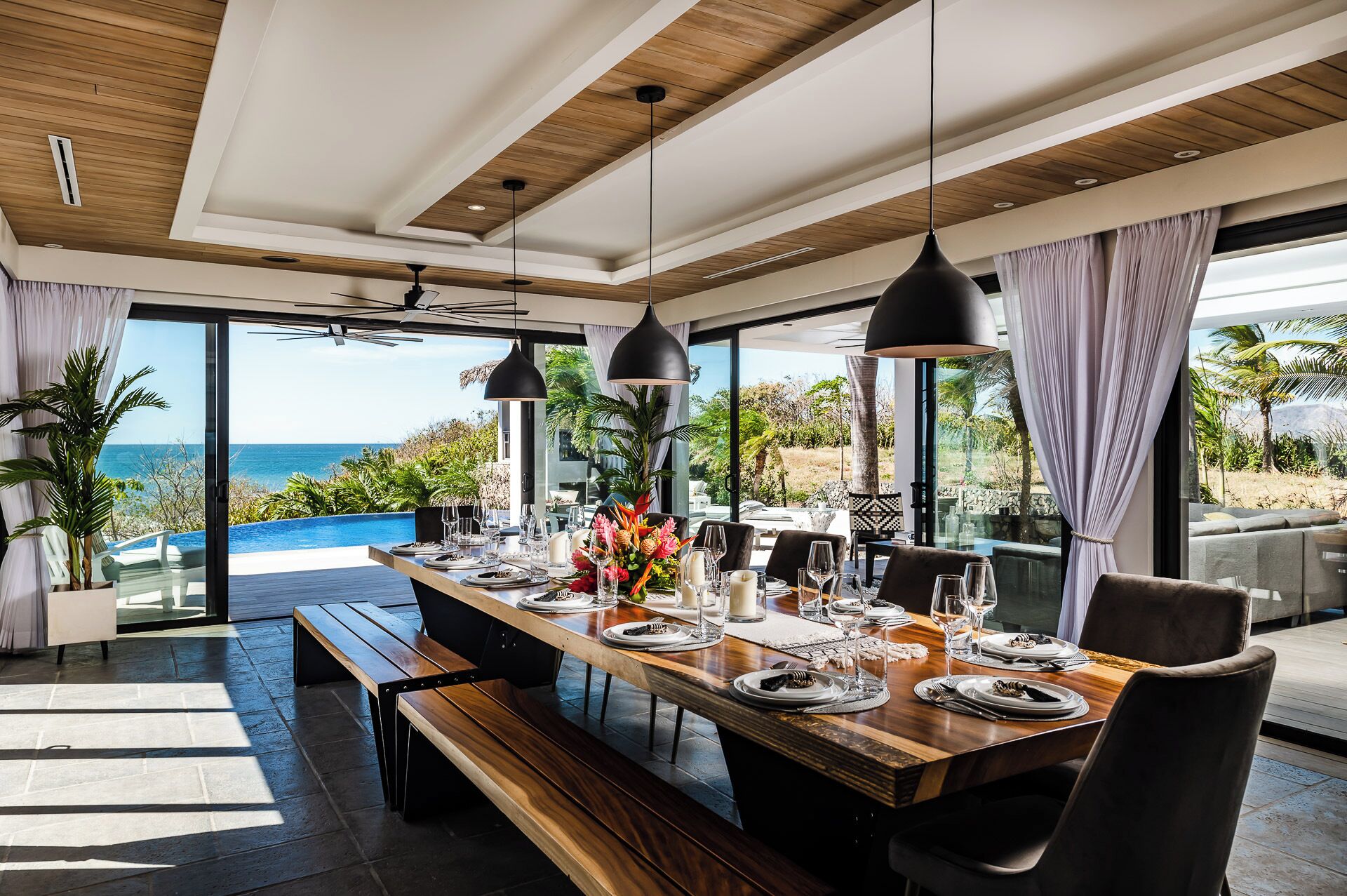 A modern dining area with a wooden table set for a meal, large windows offering a view of the ocean, and a ceiling with wooden beams.