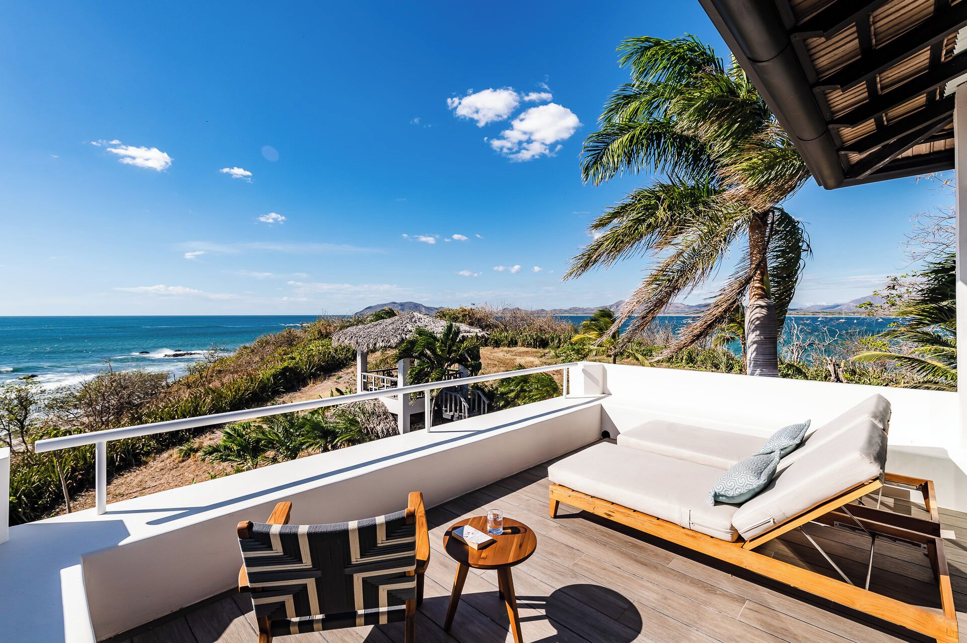 A balcony with a white sofa, a wooden chair, and a small table with a glass of water.