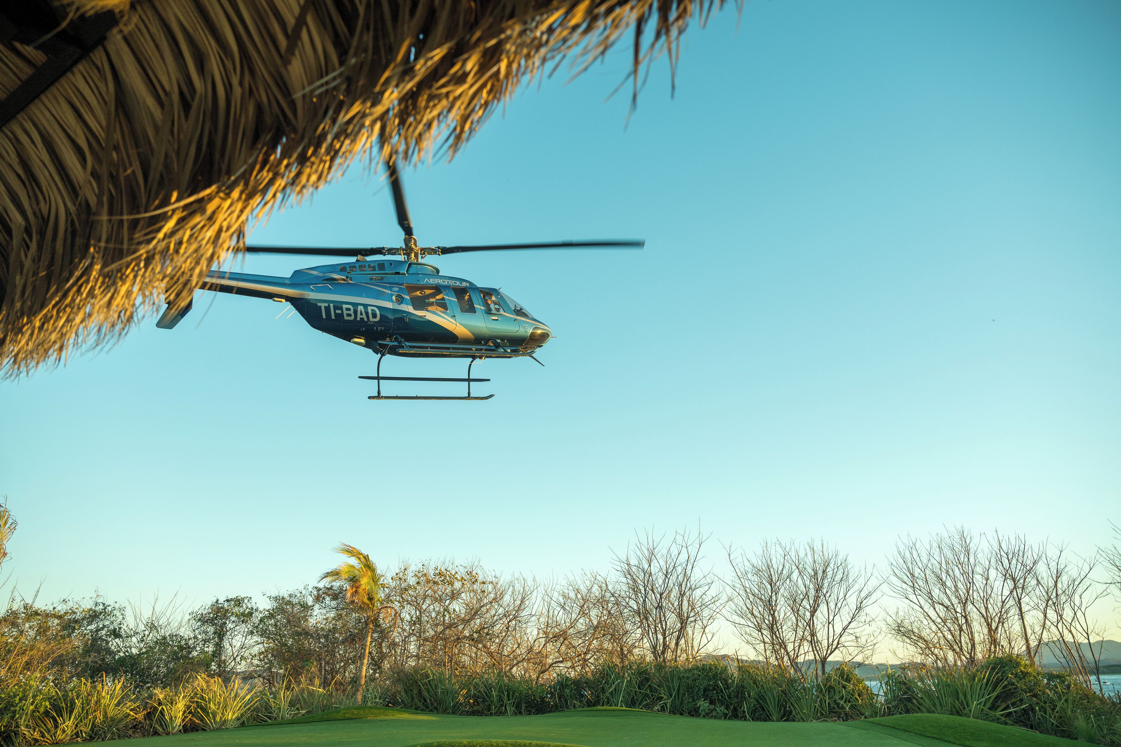 A helicopter with "TI-BAD" on its side flying over a golf course with a clear sky.