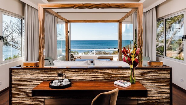 Ein Zimmer mit Holzboden, einem Bett, einem Esstisch mit einer Tablett voller Tassen und einem Buch, und Blick aufs Meer.