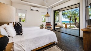 Ein modernes Schlafzimmer mit einem großen Bett, einem Deckenventilator und Blick auf den Strand durch eine Schiebetür.