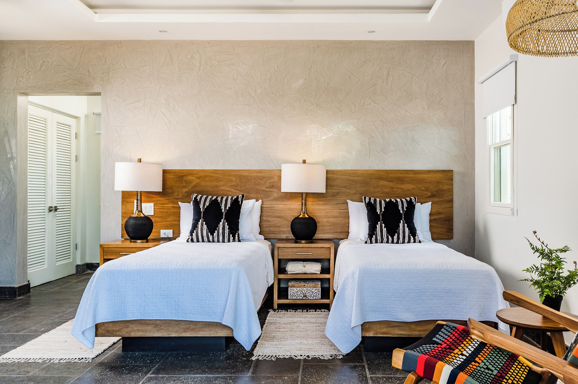 A hotel room with two beds, a wooden headboard, bedside lamps, a woven rug, a small wooden table, and a potted plant.