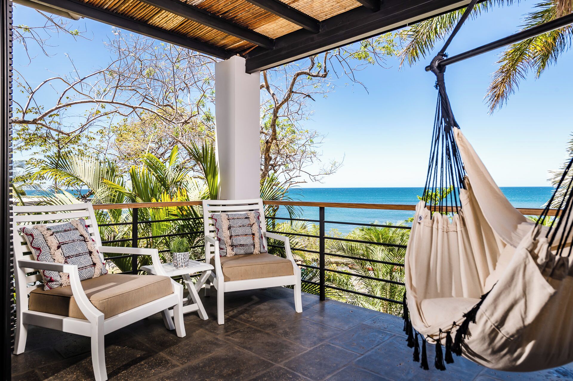 A balcony with two white chairs, a beige hammock, and a view of the ocean.
