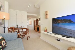 Uma sala de estar com sofá, televisão na parede exibindo uma cena de praia, área de jantar com mesa e cadeiras, e cozinha ao fundo.