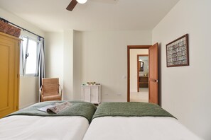 Quarto com duas camas, uma cadeira, um pequeno armário e uma porta que dá para outro cômodo.