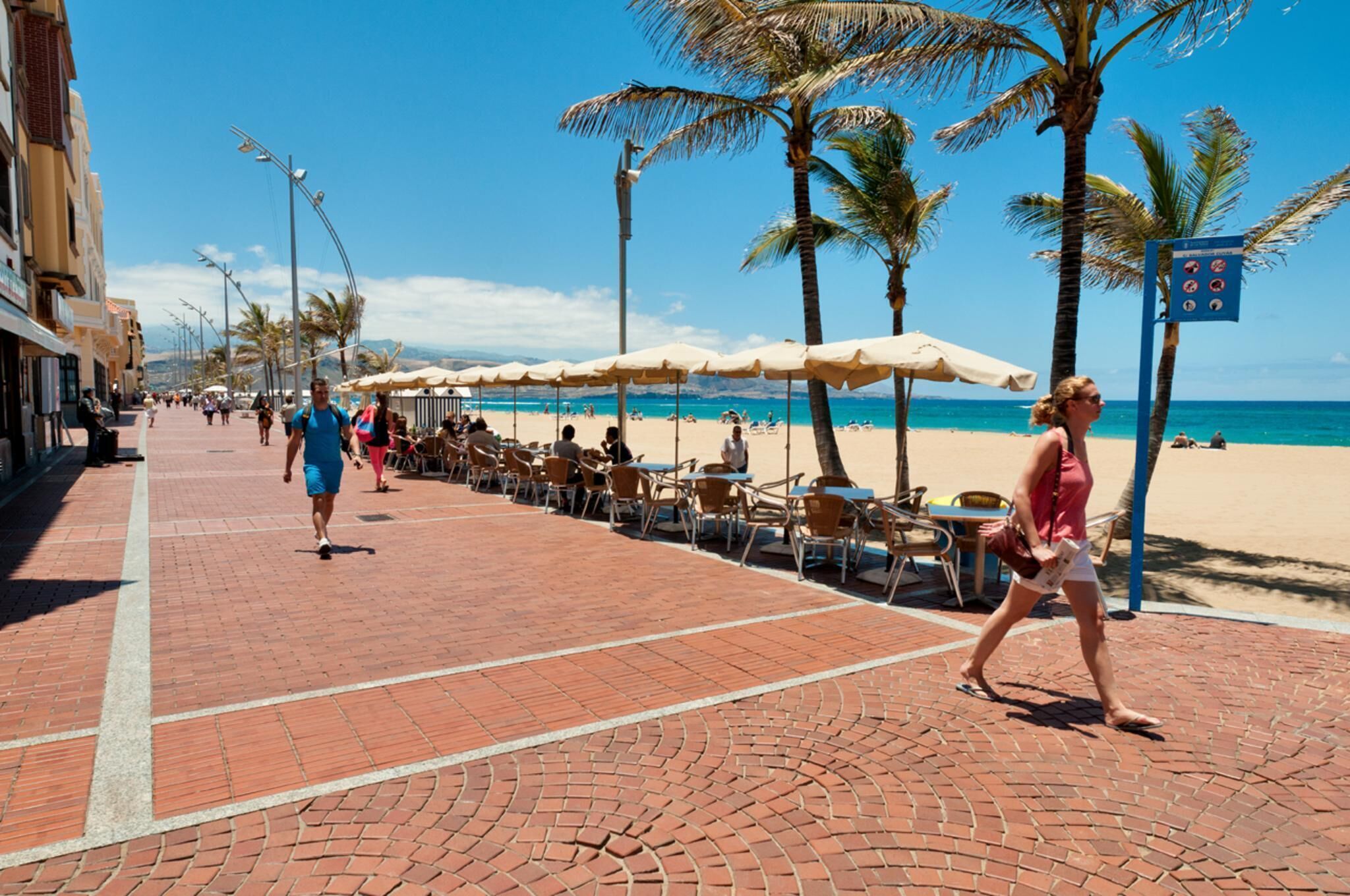 Un camino pavimentado con mesas al aire libre y palmeras, que lleva a una playa.