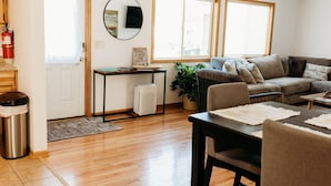 A cozy living room with a sofa, a coffee table, and a dining table. There is a plant, a small table with a picture frame, and a fire extinguisher near the door.