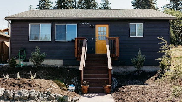 A two-story house with a dark exterior, a yellow front door, and a wooden staircase leading to the entrance.