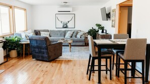 A modern living room with a grey sofa, a patterned armchair, a wooden coffee table, and a dining area with a table and chairs.