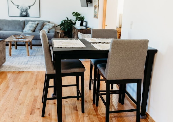 Een eetgedeelte met een zwarte tafel en vier stoelen, een houten vloer en een grijze bank op de achtergrond.