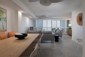 A modern living room with a dining area, a sofa set, a ceiling fan, and large windows with blinds.
