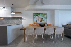 A modern kitchen with a central island, pendant lighting, and a dining area with wicker chairs and a painting.