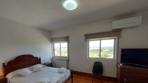 Quarto com cama de madeira, televisão, escrivaninha e duas janelas com vista para o campo.