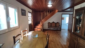Uma sala de jantar com uma mesa e cadeiras de madeira, uma escada que leva a um andar superior e vista para uma cozinha.