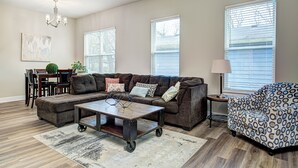 Una sala de estar con un sofá modular, una mesa de centro, un sillón estampado, una zona de comedor con mesa y sillas, y ventanas amplias con persianas.