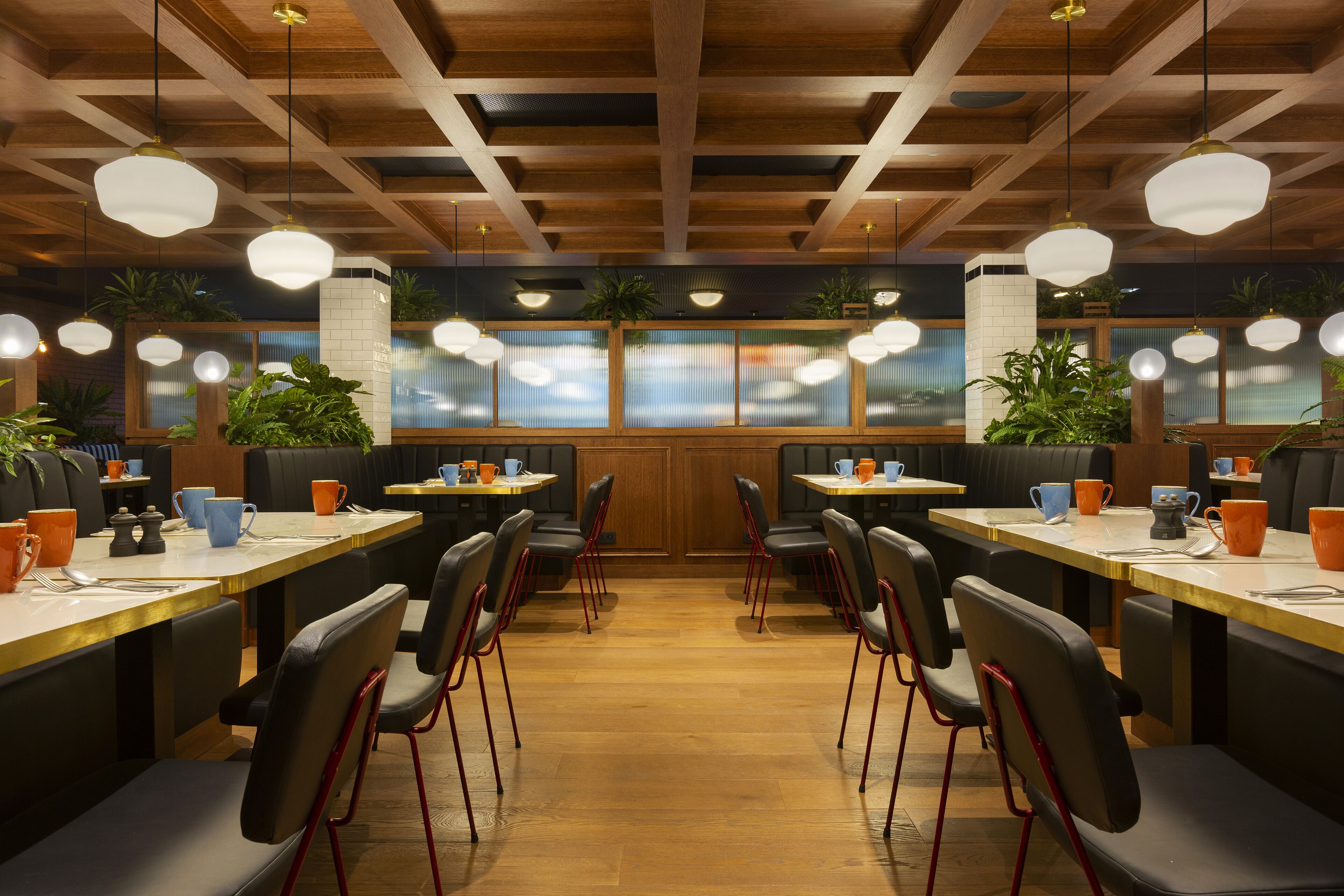 Un restaurant avec des poutres en bois au plafond, des suspensions rondes et des chaises noires aux pieds rouges. Les tables sont décorées de tasses et de soucoupes orange et bleues.