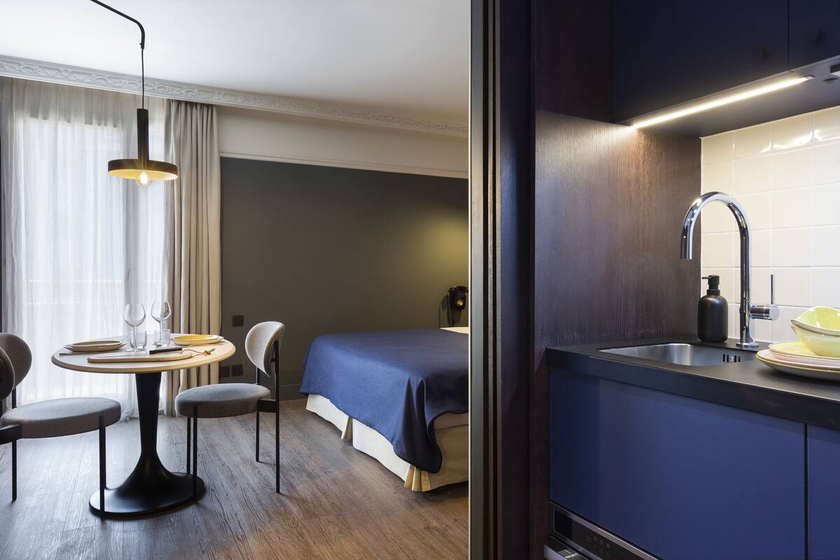 Een moderne hotelkamer met een zithoek, een bed met een blauwe spreideken, een ingebouwd wastafelmeubel en een hanglamp boven de tafel.