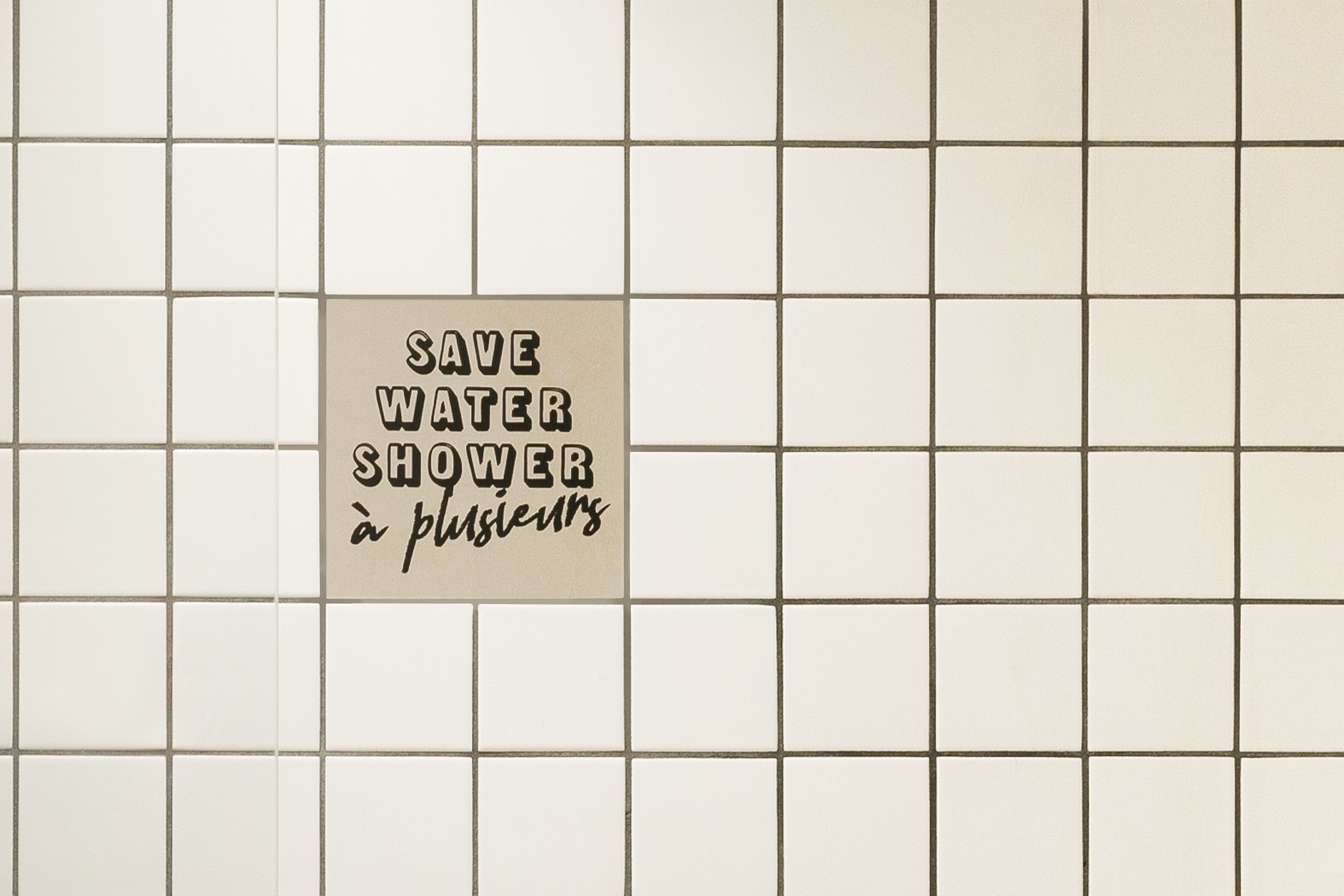 Pared de azulejos con un cartel que dice "SAVE WATER SHOWER à plusieurs". 