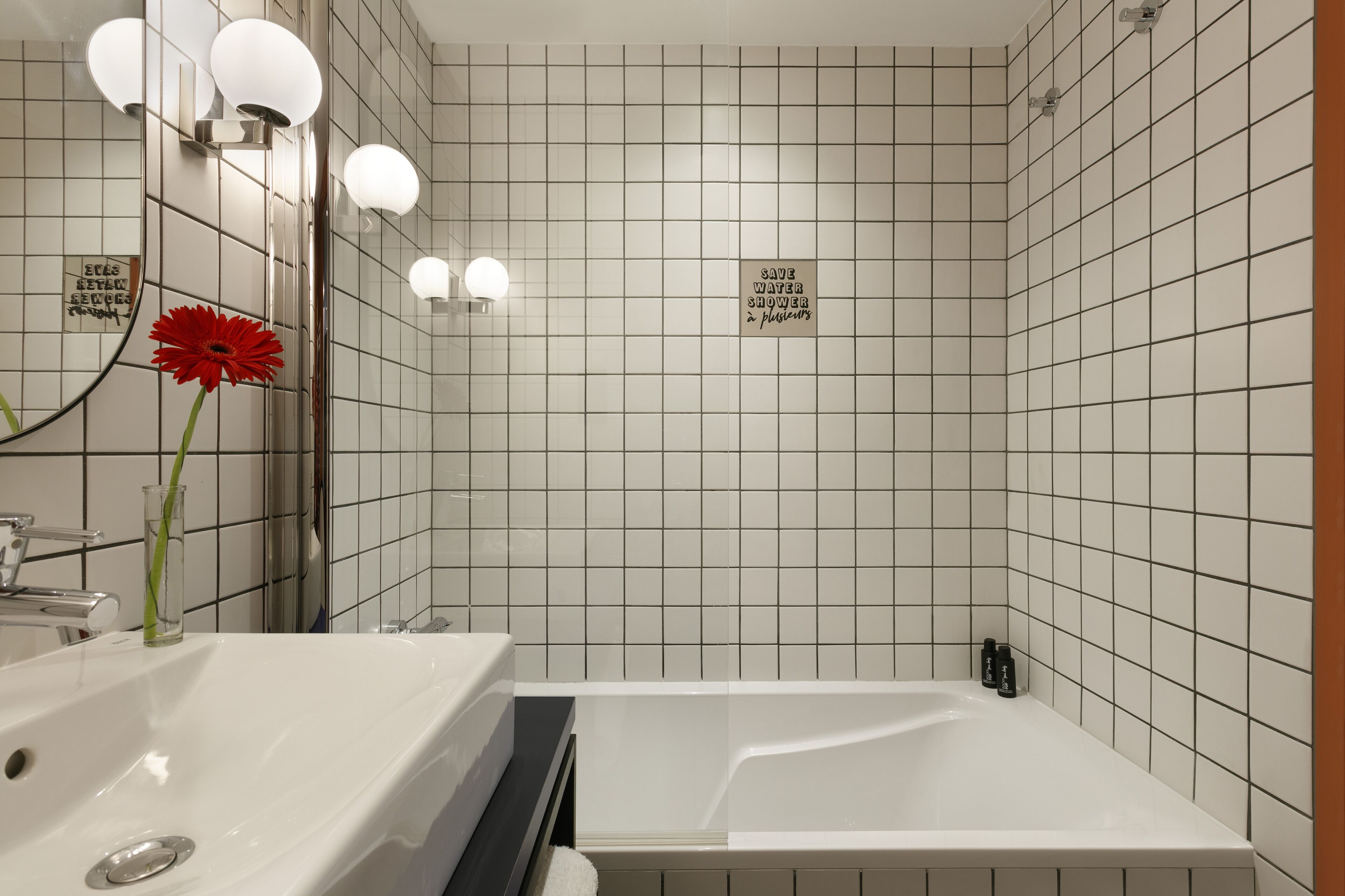 A modern bathroom with a white sink, a mirror, and a red flower. There is a sign that says "SAVE WATER SHOWER."
