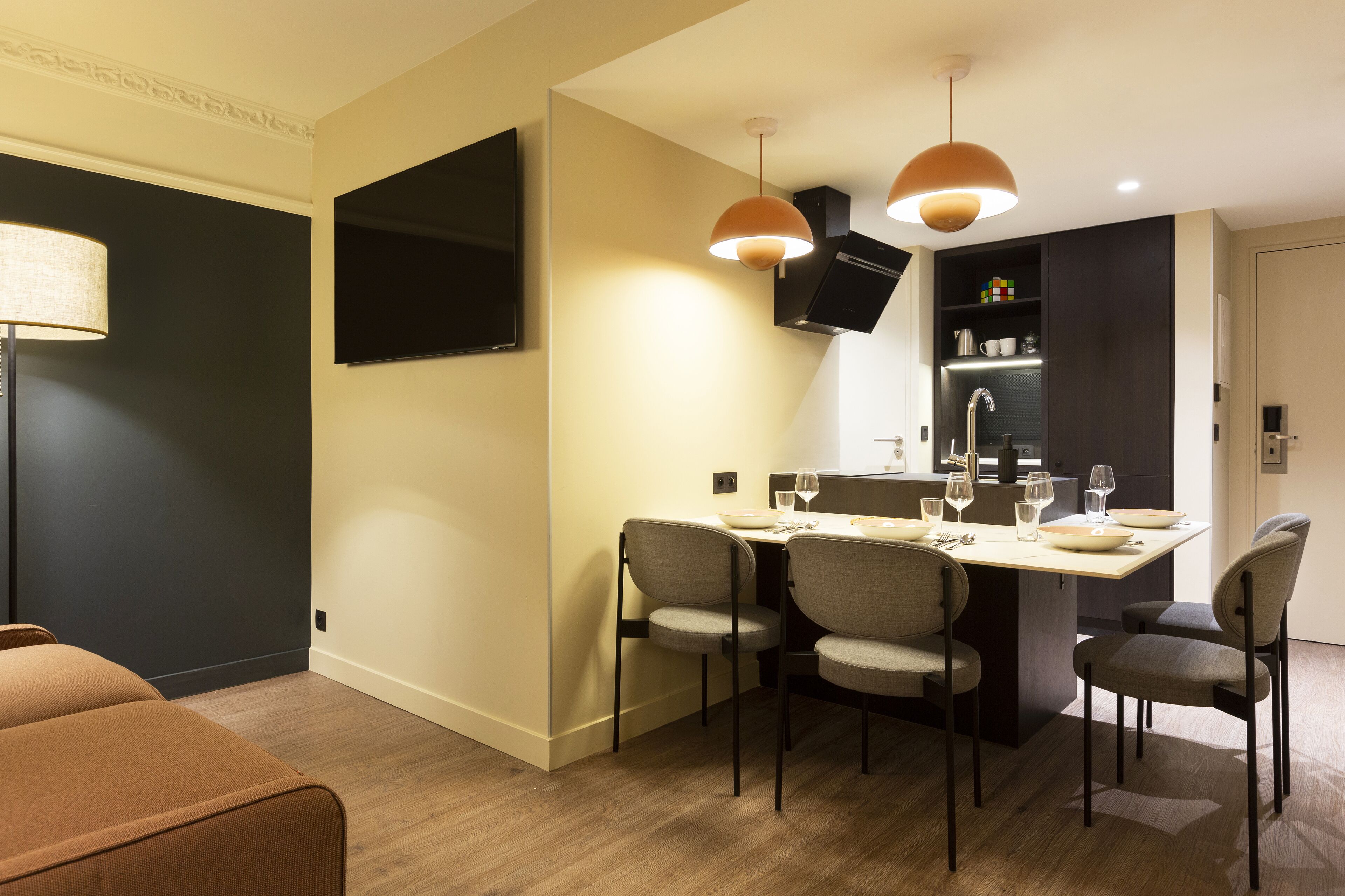 A modern dining area with a table set for four, a flat-screen TV mounted on the wall, and a kitchen area with a refrigerator and microwave.