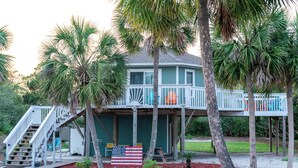 A two-story house with a porch, a flag, and palm trees. - Beach Joy! GOLF CART included! 2-min BEACH walk, Sleeps 6!! (St. George Island)