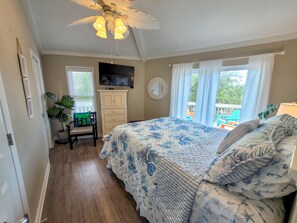 A bedroom with a bed, a ceiling fan, a TV, a chair, a plant, and a window with curtains.