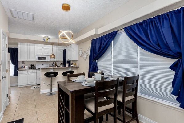 A dining area with a wooden table set for four, a kitchen in the background, and blue curtains on the windows. - 5 minutes to Cherry point! Family-sized townhouse with fireplace & balcony (Havelock)