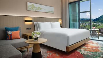 Una habitación de hotel moderna con una cama grande, un sofá, una mesita con un jarrón de flores y un amplio ventanal con vistas a la montaña.