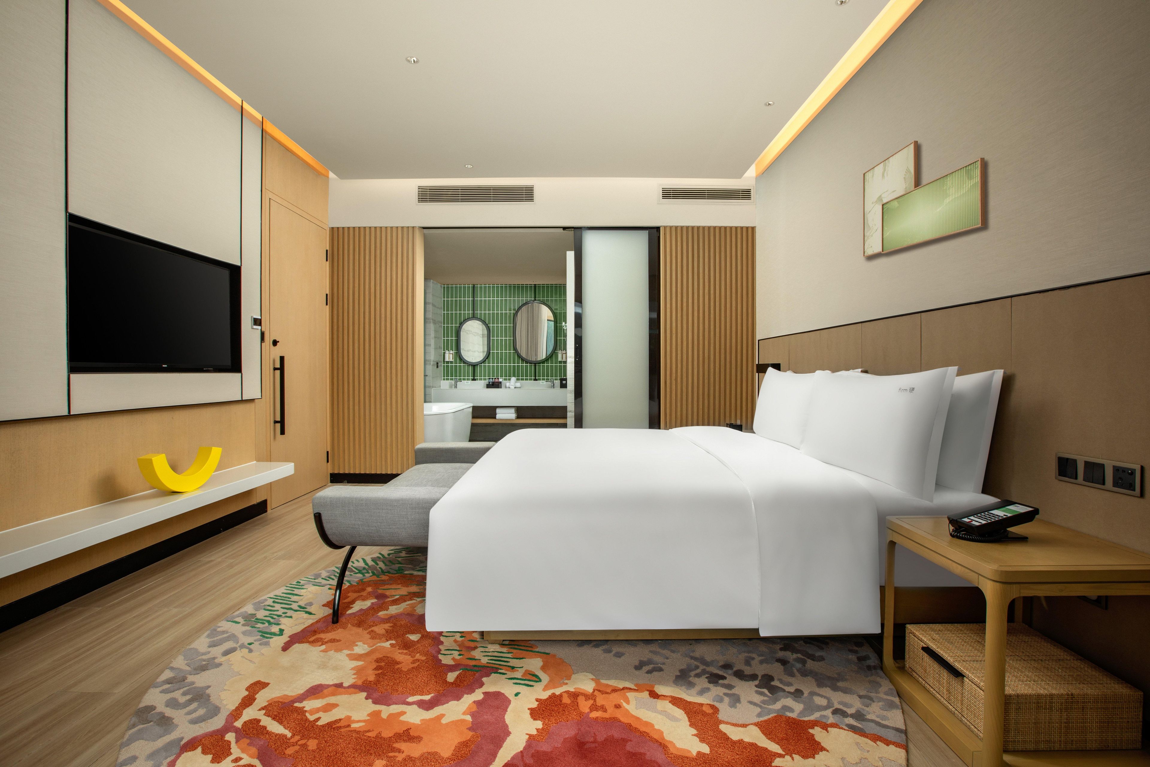 A hotel room with a bed, a TV, a bedside table, a mirror, and a bathroom visible through an open door.