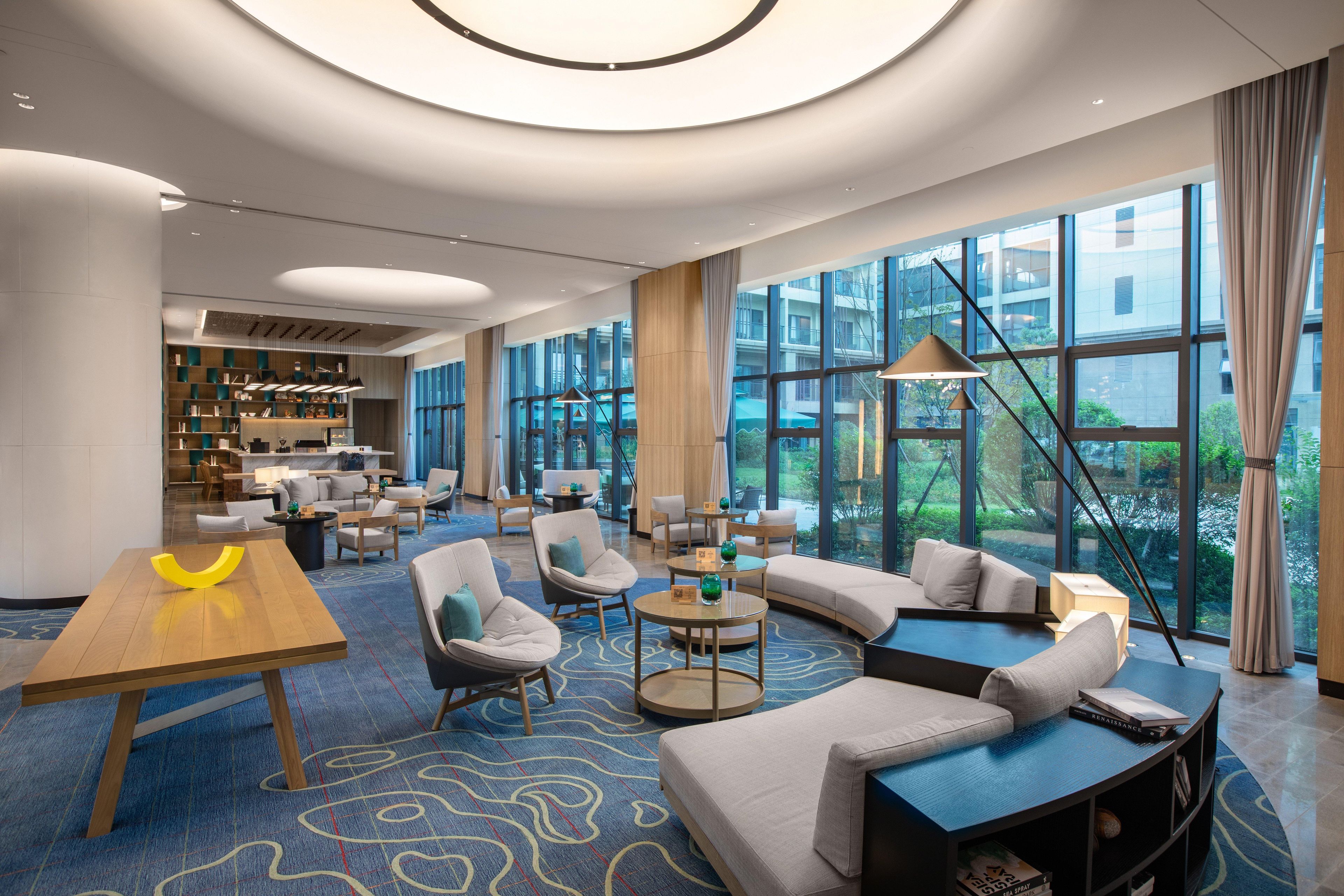 A modern hotel lobby with a curved sofa, round tables, and a large window offering a view of the outdoors.