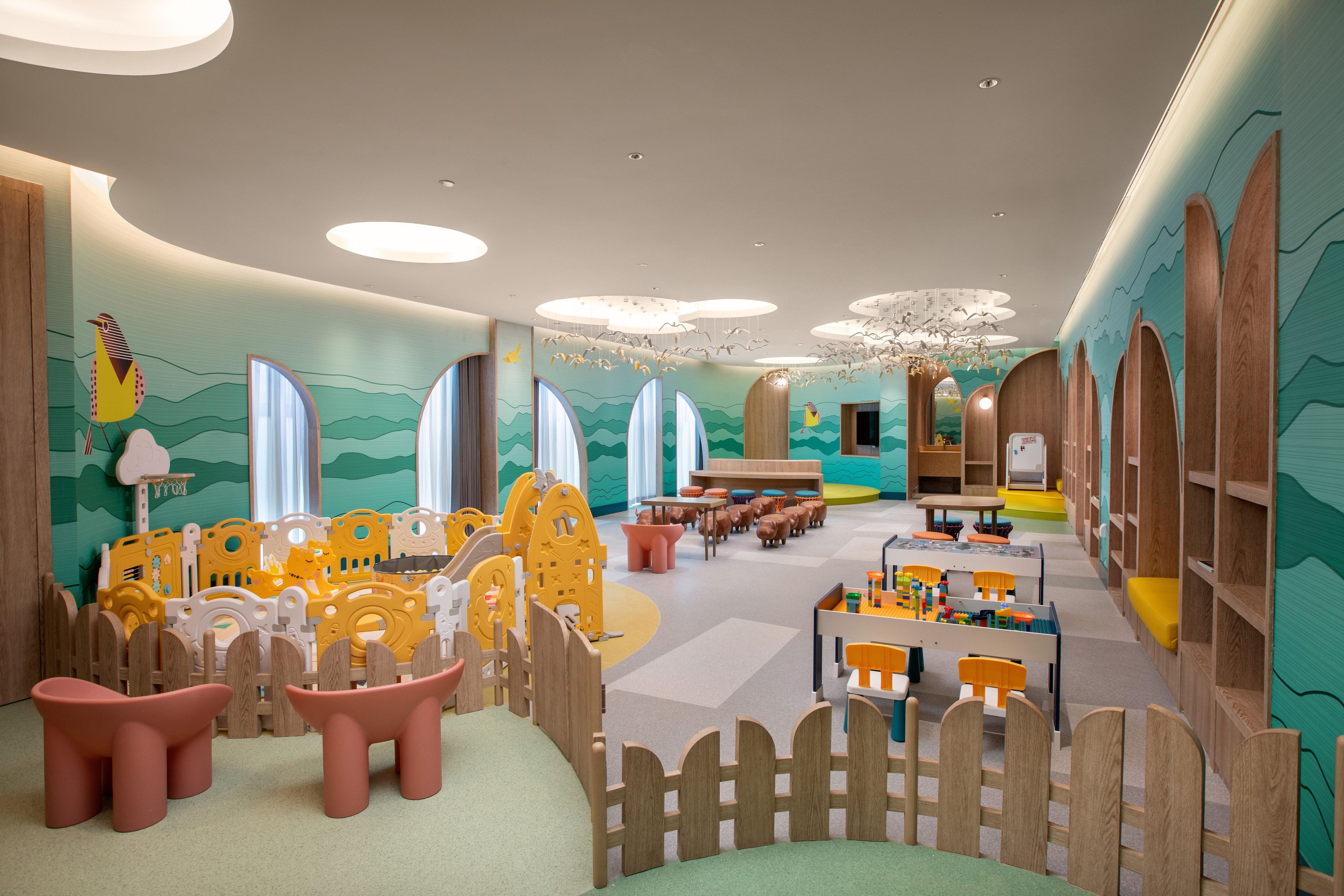 A children's playroom with a wooden playpen, tables, chairs, and a mural on the walls.