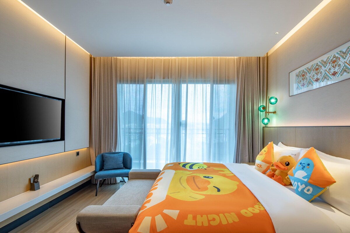Habitación de hotel con una cama grande, televisión montada en la pared, zona de estar con una silla y una ventana con cortinas.