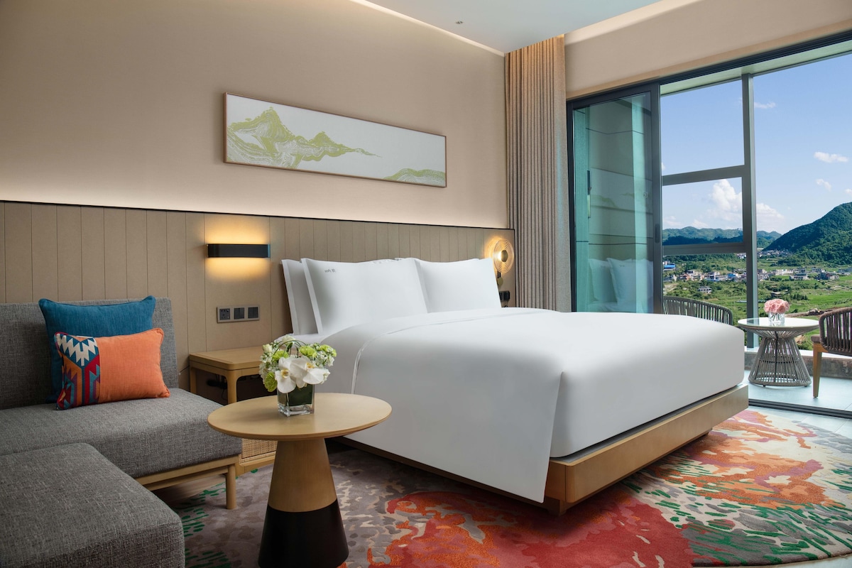 A modern hotel room with a large bed, a sofa, a small table with a vase of flowers, and a large window offering a mountain view.