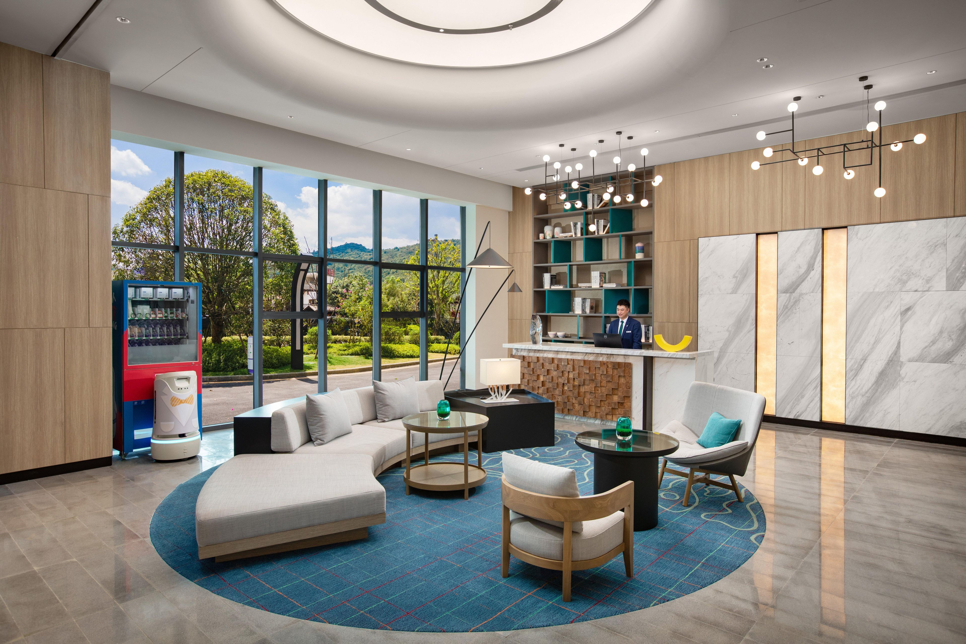 A modern hotel lobby with a curved sofa, round tables, a bar area, and a large window offering a view of greenery.