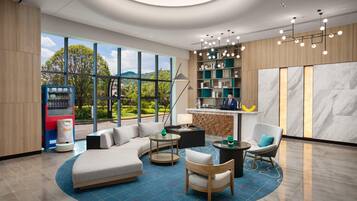 A modern hotel lobby with a curved sofa, round tables, a bar area, and a large window offering a view of greenery.