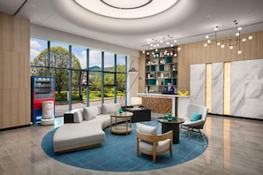 A modern hotel lobby with a curved sofa, round tables, a bar area, and a large window offering a view of greenery.