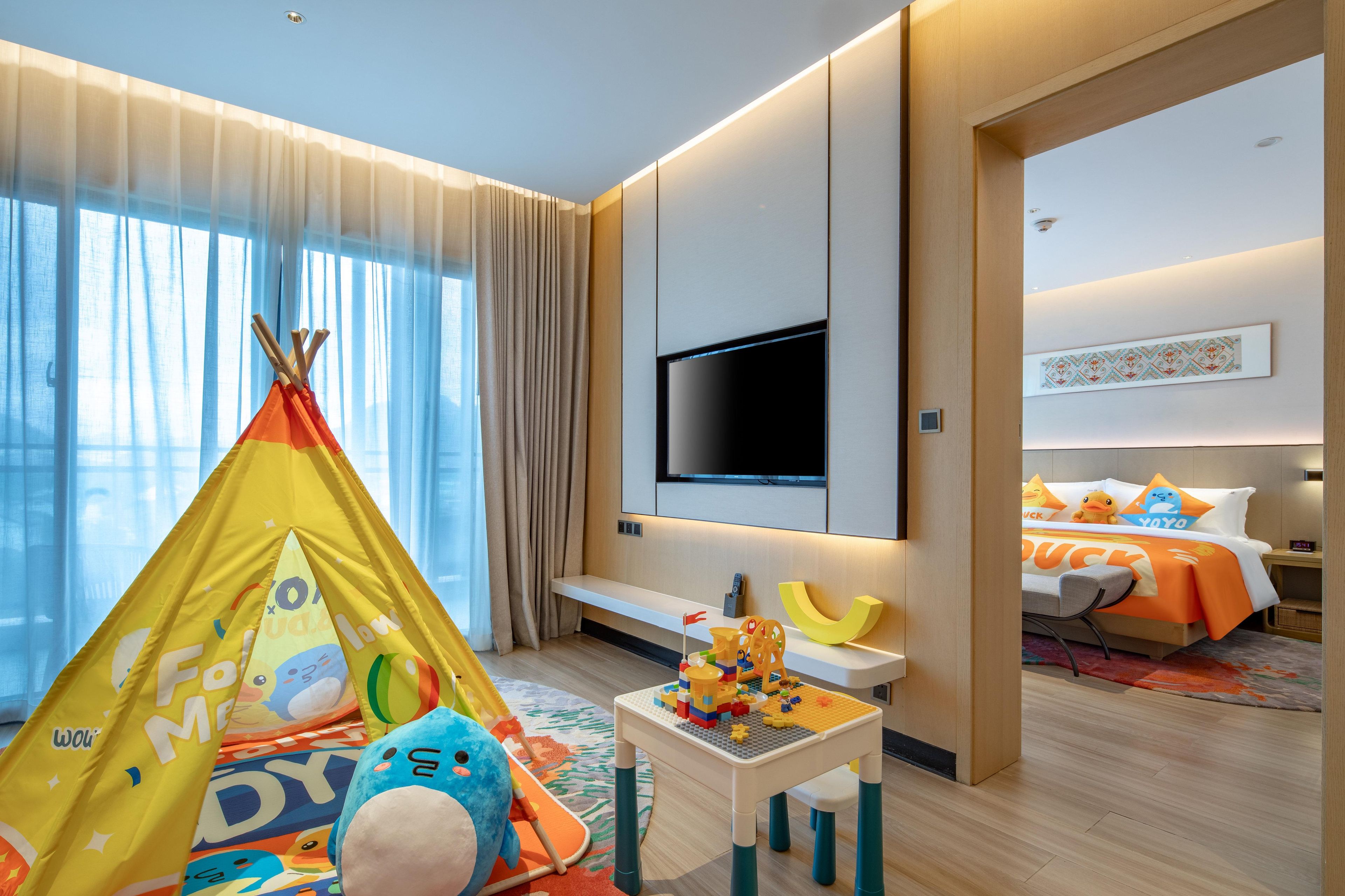 Habitación de hotel con zona de juegos, cama con edredón de tema pato, televisor de pantalla plana y ventana grande con cortinas.
