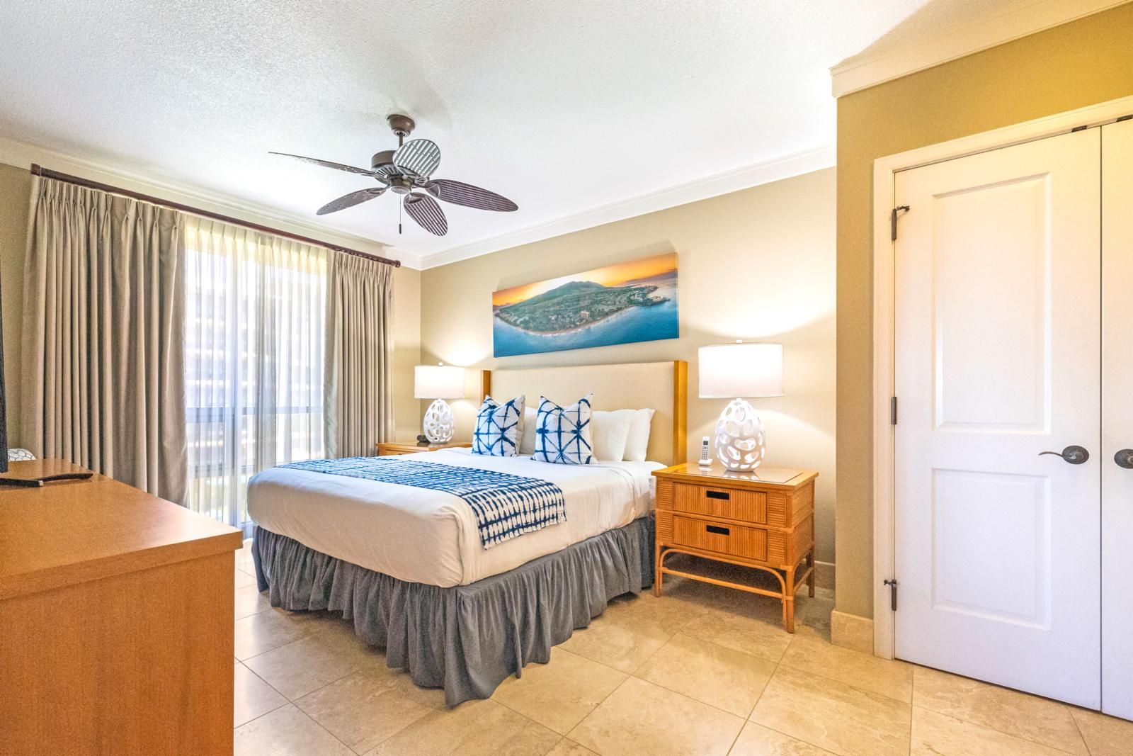 A hotel room with a bed, a ceiling fan, a painting of a landscape, a nightstand with a lamp, and a door.