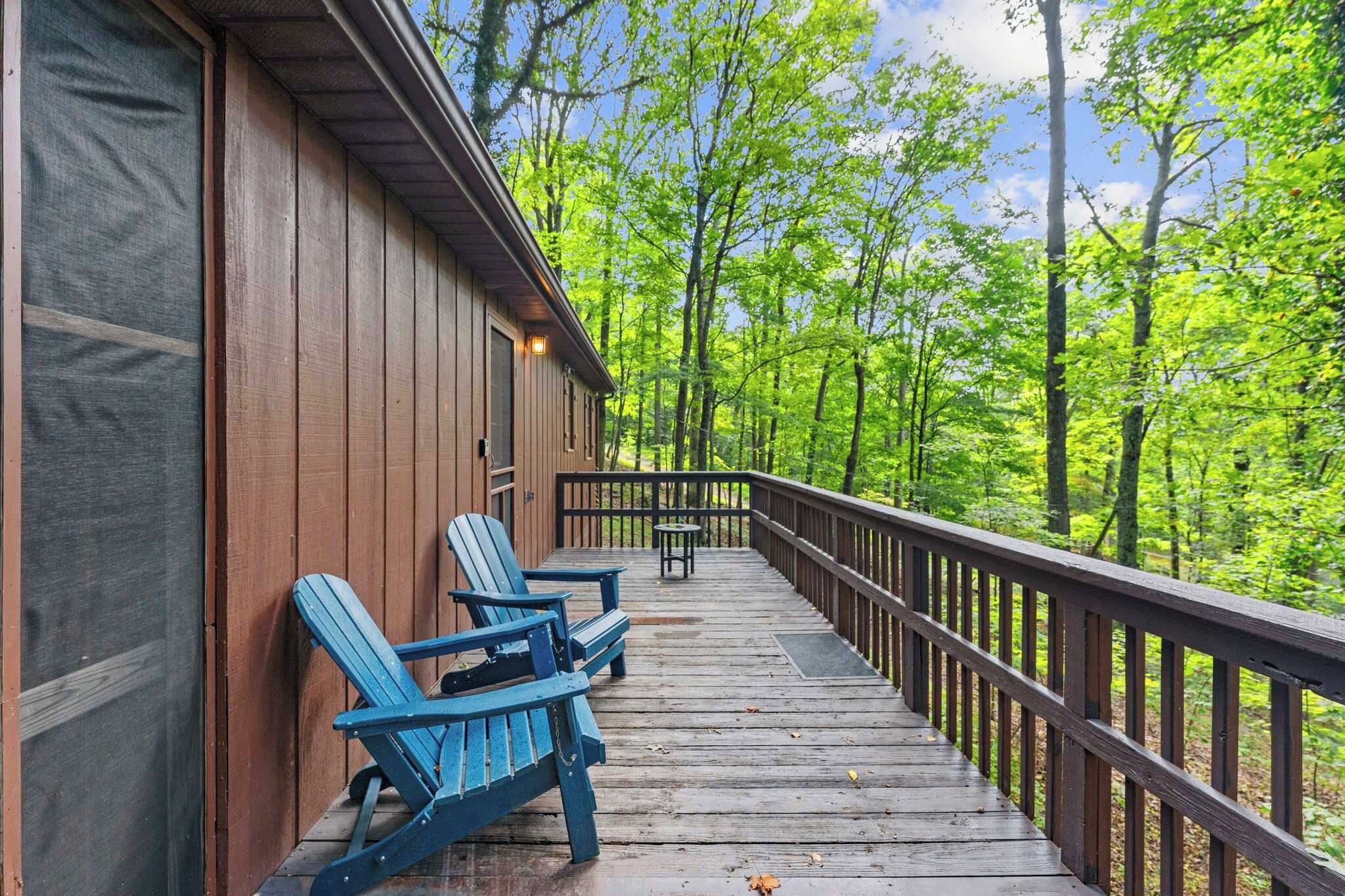 A wooden deck with Adirondack chairs and a table, surrounded by a forest.