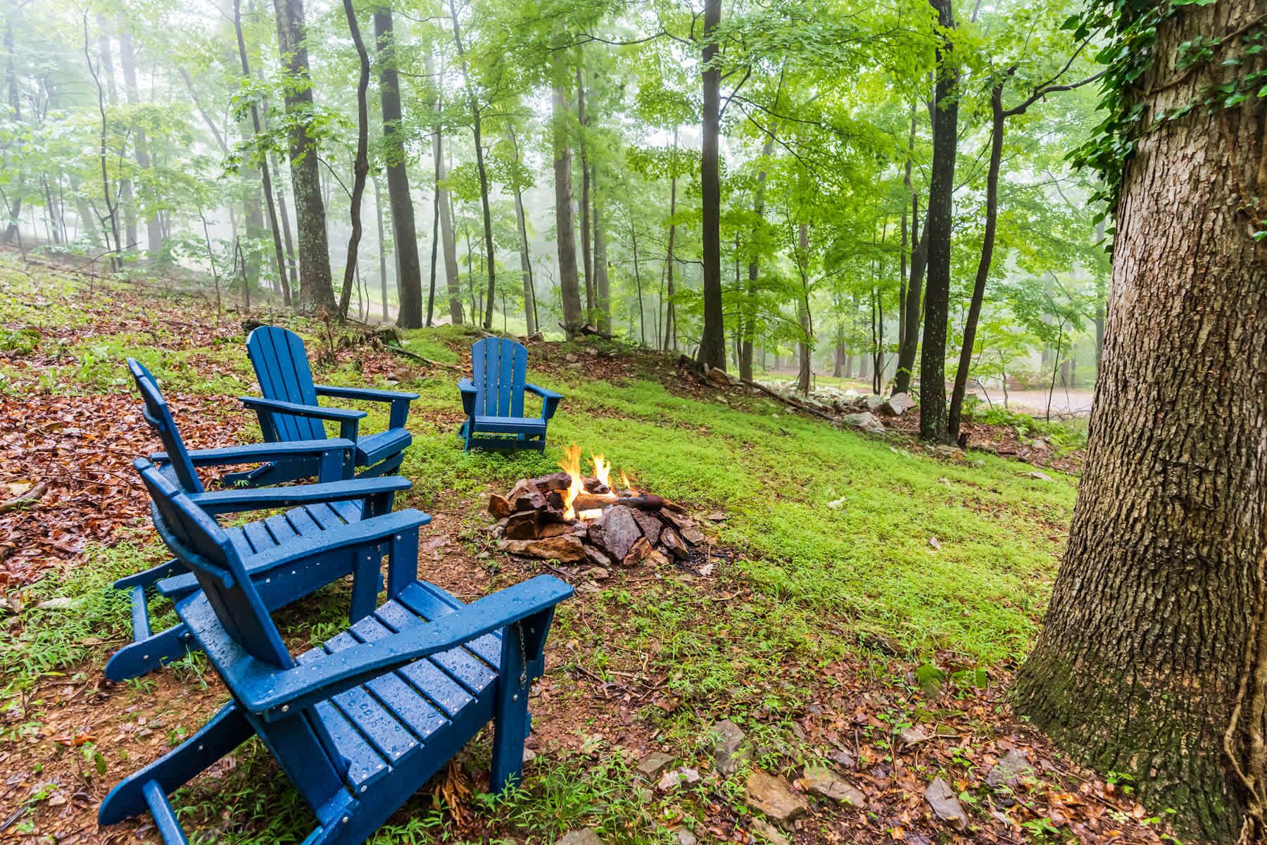 A forest with three blue Adirondack chairs and a fire pit.