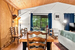 Una mesa de comedor de madera con sillas, un sofá y una televisión en una habitación con pared blanca y techo de madera.