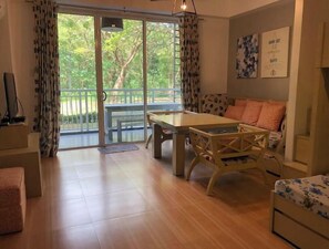 A spacious living room with a wooden floor, a sofa, a dining table, and a balcony with a view of greenery.
