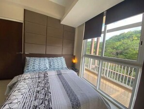 Une chambre à coucher avec un grand lit, une tête de lit dotée de compartiments de rangement, une fenêtre donnant sur un paysage verdoyant et une table de chevet avec une lampe.