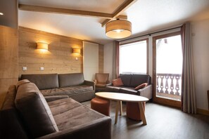 Uma sala de estar moderna com um sofá, poltronas e uma mesa de centro. Há duas luminárias fixadas na parede e uma luz no teto. O aposento tem grandes janelas com cortinas e vista para o exterior.