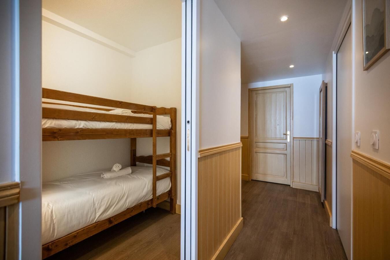 Un lit superposé dans un couloir, avec un plancher en bois et une porte visible au fond.